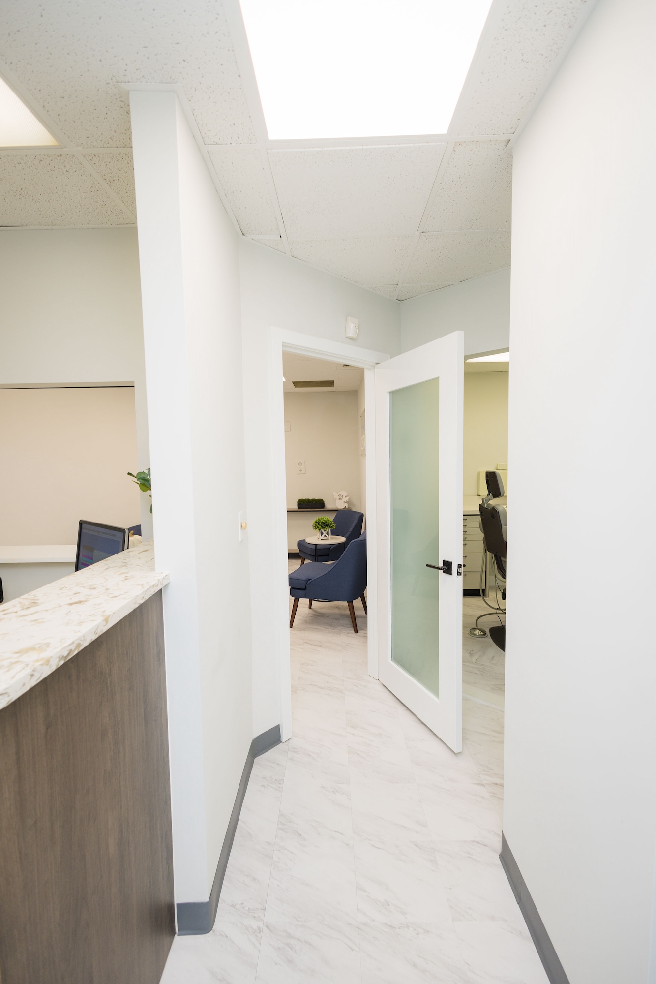 The image depicts an interior office space with a hallway leading to a reception area, featuring a modern design with white walls and a light-colored floor.