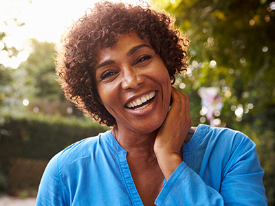 A woman with a radiant smile, wearing a blue top, stands confidently against a sunny backdrop.