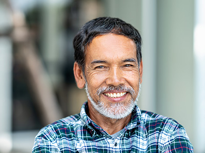 The image is a portrait of an elderly man with a beard and mustache, wearing a plaid shirt, smiling at the camera.