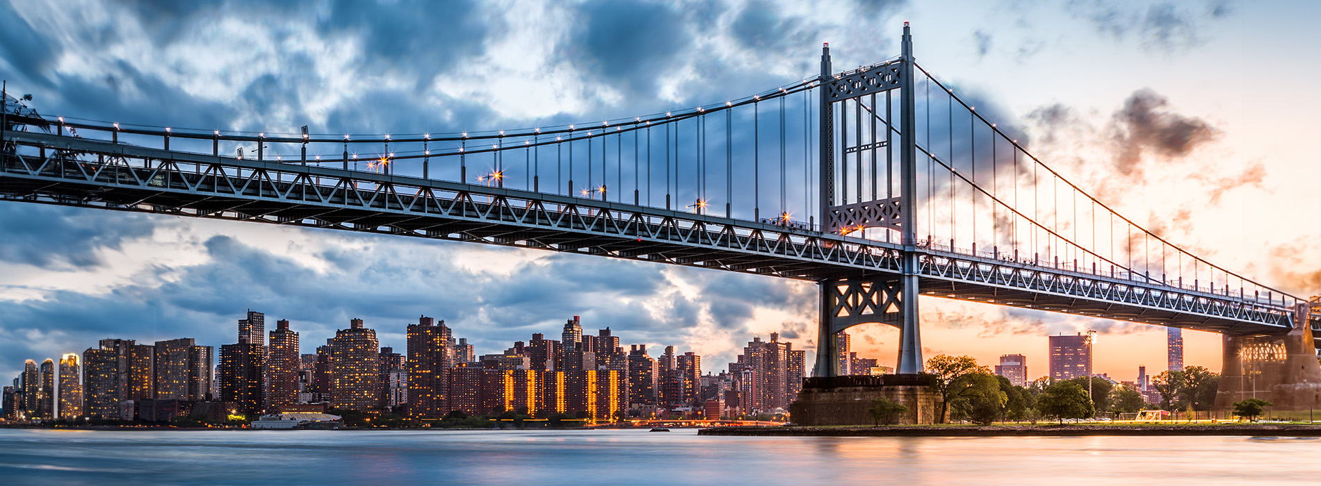 A photograph of a city skyline at sunset with a long suspension bridge in the foreground, featuring a distinctive design and illuminated against a cloudy sky.