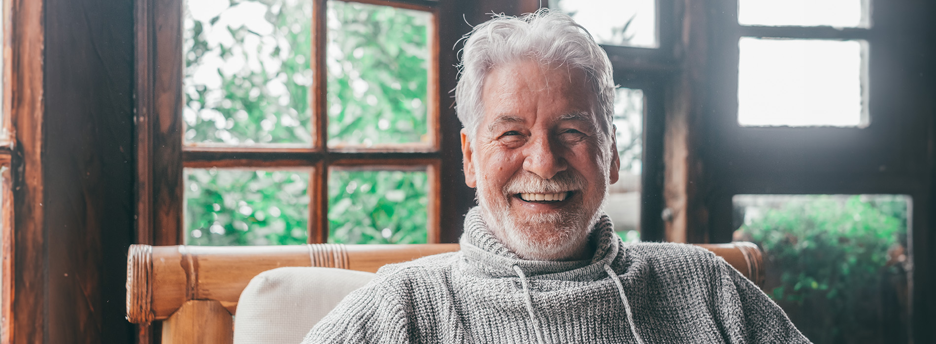 The image shows an elderly man with white hair, smiling and looking directly at the camera. He is seated in a room with a window behind him, wearing a sweater and sitting on a couch or chair.