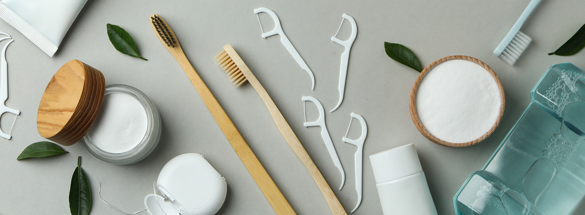 An assortment of skincare products and tools on a light grey background, with a wooden table and a green leafy plant in the corner.