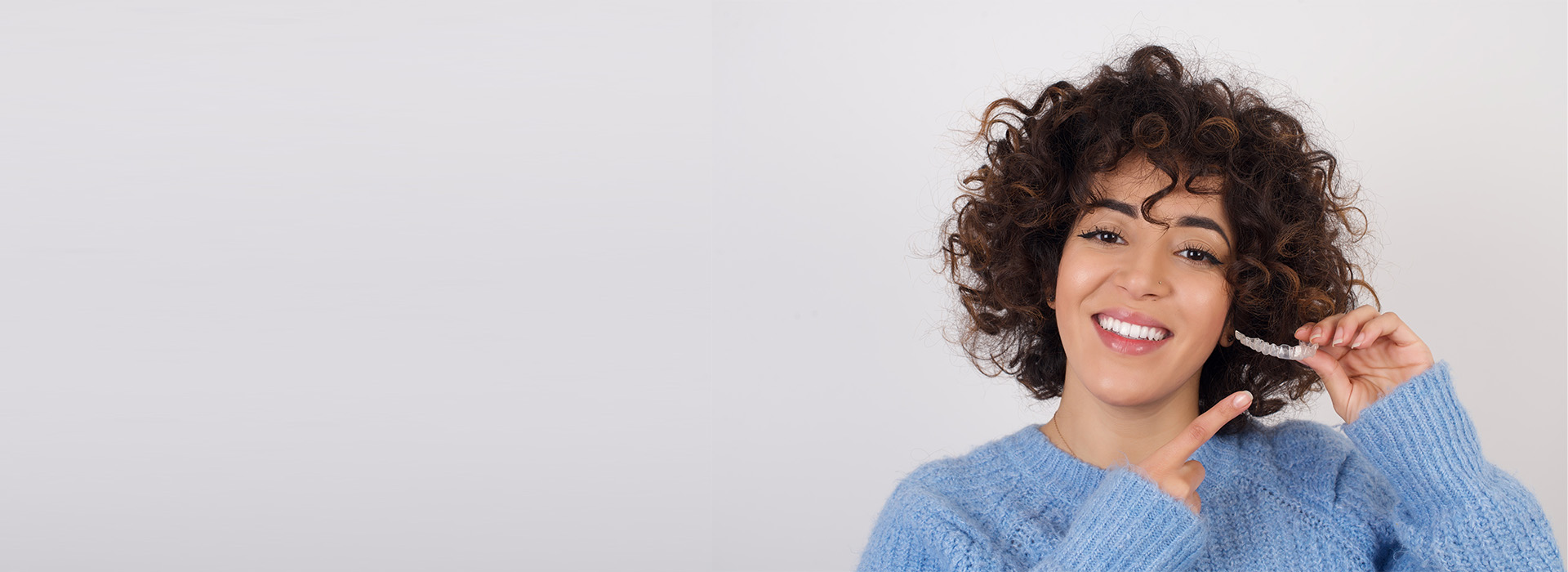 A woman with curly hair is smiling and holding a phone, set against a neutral background.