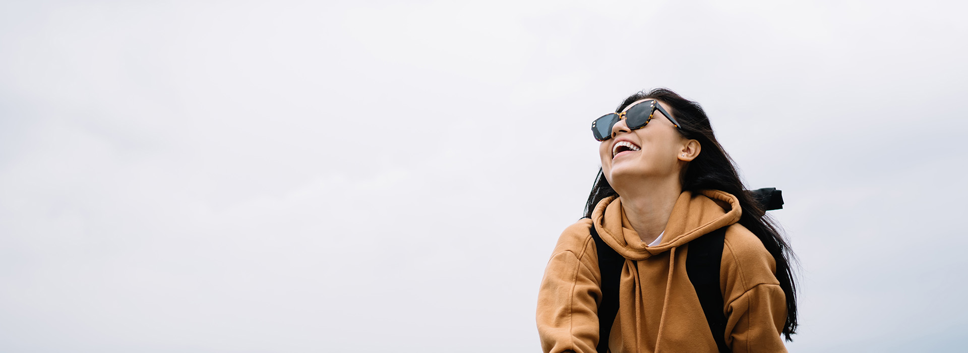 The image depicts a person standing outdoors, wearing sunglasses and a backpack, with their hair tied back, smiling at the camera. They are dressed in casual attire and appear to be enjoying a pleasant day.