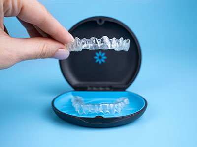 The image shows a hand holding an open dental impression tray with a blue background, displaying a set of clear plastic teeth inserts.