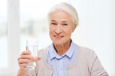 The image features an elderly woman holding a glass of water in her hand, smiling at the camera.