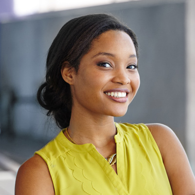 The image shows a smiling woman with dark hair, wearing a yellow top and standing against a neutral background.