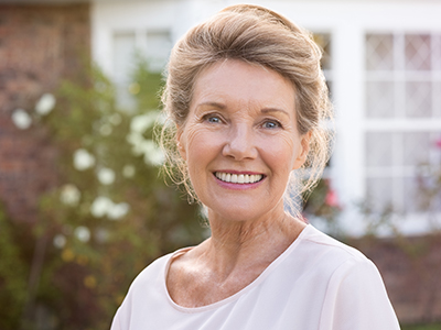 The image shows a smiling older woman with short hair, wearing a light-colored top and standing in front of a house with a brick wall.