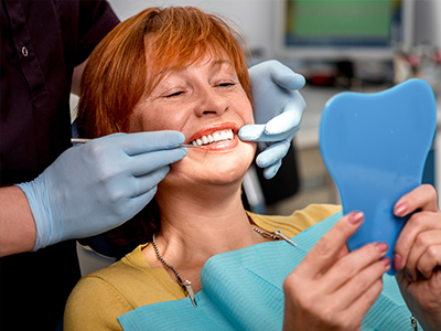 A woman sitting in a dental chair, smiling broadly while looking at her reflection in a mirror, with a dental professional adjusting her teeth.