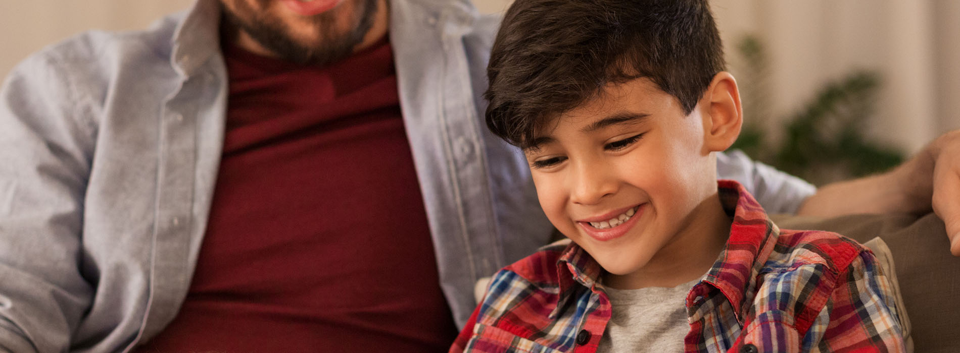 A man and a boy sharing a moment on a couch, with the man smiling at the camera and the boy looking at a screen.