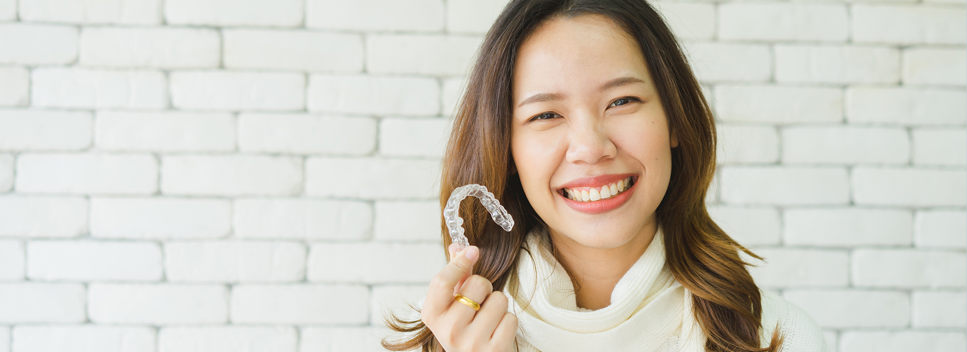 A woman with a radiant smile is holding a ring, showcasing it against her chest. She has dark hair and is wearing a light-colored top. The background features a brick wall.