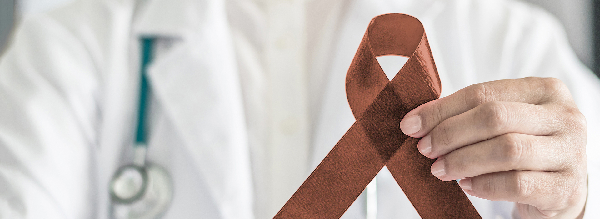 The image shows a hand holding a brown ribbon with a white cross, which is commonly associated with the symbol of medical aid. In the background, there is a person wearing a white lab coat, suggesting they are a healthcare professional.