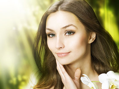 The image is a professional portrait of a woman with light skin and dark hair, wearing makeup, against a soft-focus background that suggests an outdoor setting.