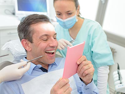 The image shows a man sitting in a dental chair, holding up a pink card with a surprised expression, while a woman wearing a surgical mask smiles and holds a tray with dental instruments.