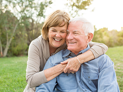 An elderly couple embracing in a park-like setting, with the man wearing glasses and both smiling.