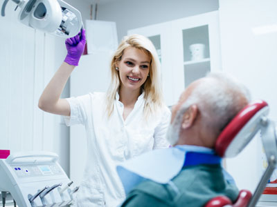 A dental professional, likely a female dentist or hygienist, is smiling and holding up a device towards an elderly man s mouth in what appears to be a dental office setting.