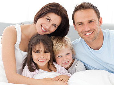 A family of four, including a man, woman, and two children, posing together in a bedroom setting with a smile.