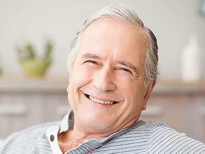 The image shows an elderly man with white hair, wearing a blue shirt and smiling broadly. He appears to be relaxed and content as he sits in a chair.