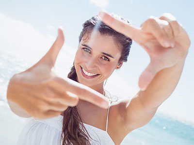 A young woman with a radiant smile takes a selfie against the backdrop of a sunny beach, capturing her joyful moment.